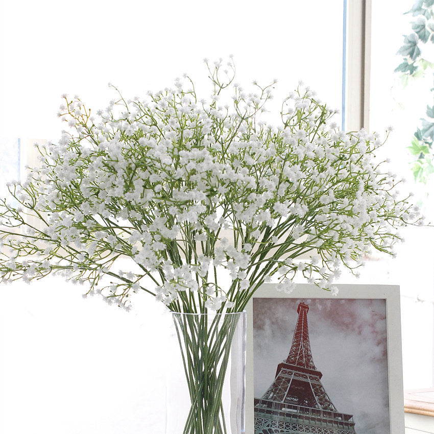 Airy white artificial gypsophila branches in a clear glass vase beside a framed Eiffel Tower photo by a window