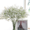 Airy white artificial gypsophila branches in a clear glass vase beside a framed Eiffel Tower photo by a window