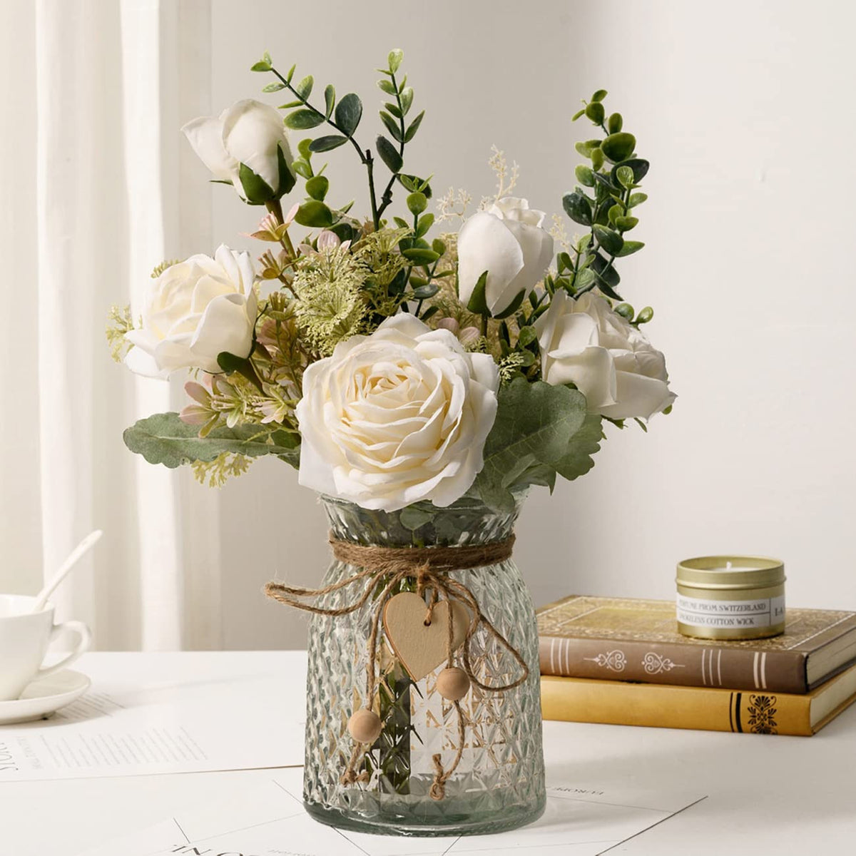 Artificial white silk rose bouquet with greenery in a textured glass vase tied with twine, beside books on a table