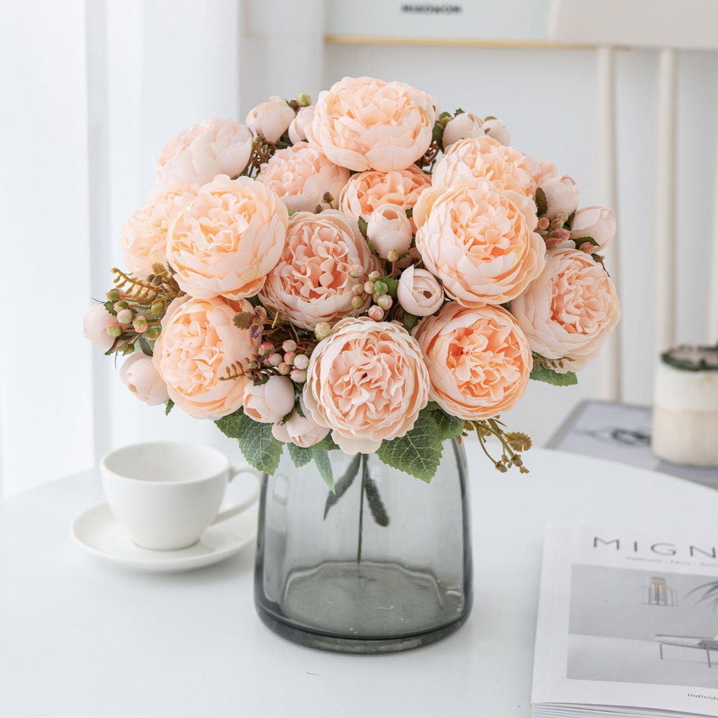 Blush pink silk peony bouquet in a smoky gray glass vase on a white table next to a teacup and saucer
