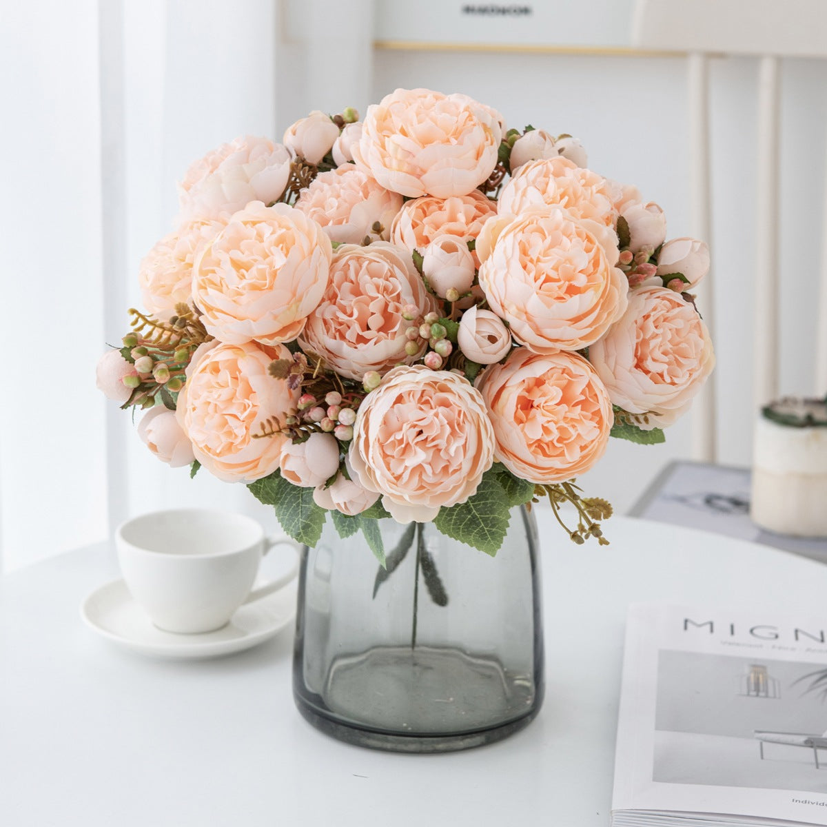 Blush pink silk peony bouquet in a smoky gray glass vase on a white table next to a teacup and saucer