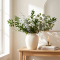 Faux eucalyptus, ruscus and baby's breath stems in a white ceramic vase on a wooden console in a sunlit room